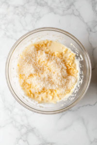 top-down-macaroni-mixed-with-milk-and-condensed-soup-in-clear-glass-bowl-on-white-marble