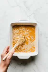 top-down-whisking-nutritional-yeast-broth-and-miso-in-white-ceramic-baking-dish-on-marble