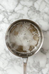 This image shows white vinegar in a stainless steel saucepan simmering with sugar and salt, photographed top-down over a white marble surface.