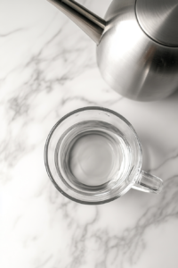 This image shows a clear glass cup filled with warm water placed on a white marble countertop. The scene is minimal, and no ingredients have been added yet.