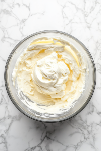 This image shows whipped cold heavy cream being folded into thickened vanilla pudding using a silver food processor bowl, all placed on a white marble countertop.