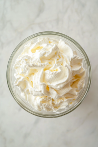 This image shows softly whipped cream with peaks forming in a transparent glass bowl placed on a white marble countertop.