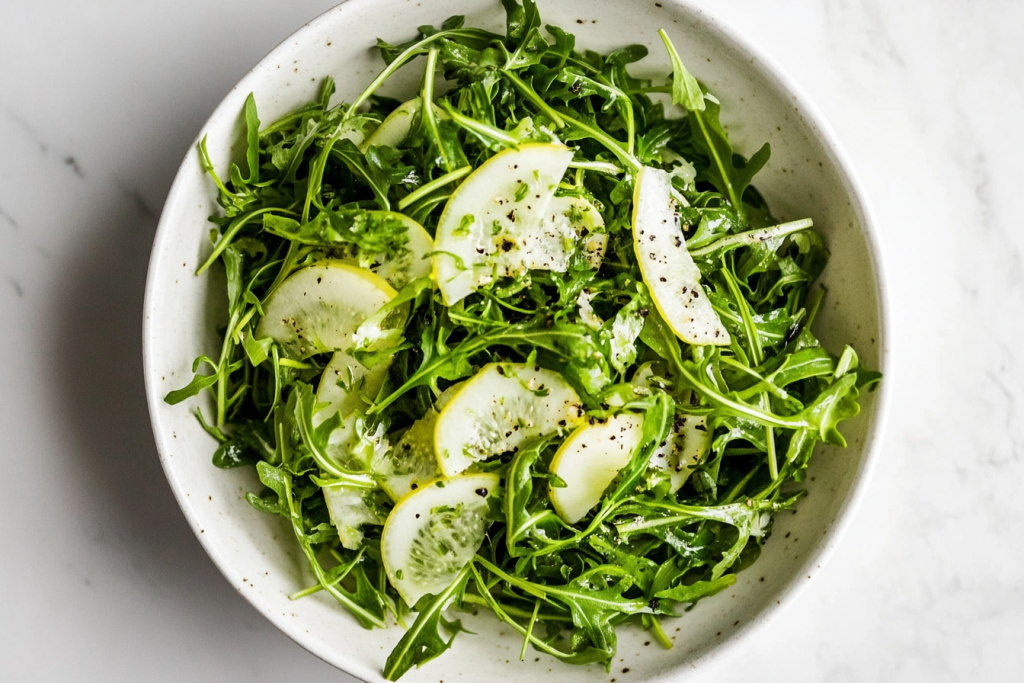 This image shows a white bowl of arugula and thinly sliced fennel, tossed with a zesty dressing made with extra-virgin olive oil, lemon zest, lemon juice, salt, and freshly ground pepper.