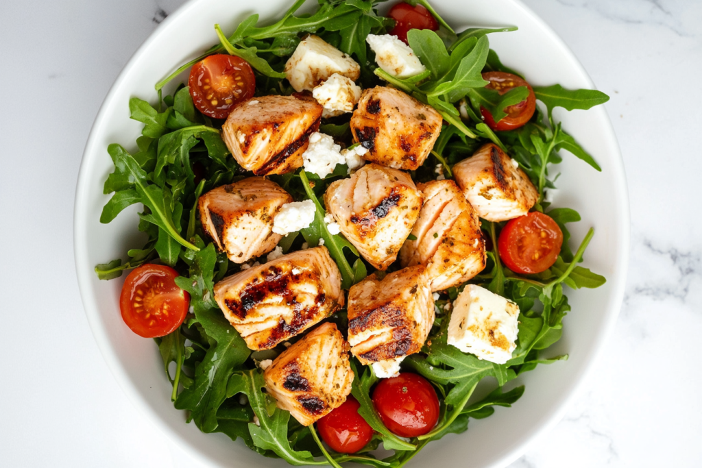 This image shows a white bowl of feta salmon salad, made with fresh spring mix greens, chopped cucumber, tomatoes, crumbled feta cheese, and perfectly cooked salmon pieces.